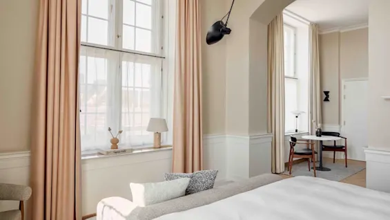 A bright room features large windows with beige curtains, a neatly made bed, a wall-mounted black lamp, and a small table with chairs. A book titled "Ingegerd Råman" is on the windowsill.