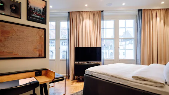 A neatly made bed faces a flat-screen TV in a softly lit room with large windows and beige curtains. A desk is adorned with framed pictures and a map.