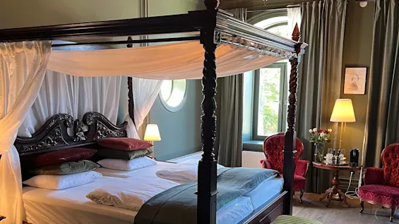 En himmelseng med hvide gardiner står centralt i et elegant rum. To røde stole, en lampe og et vindue med gardiner giver en hyggelig atmosfære.