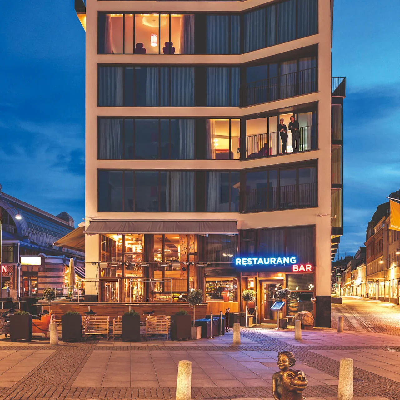 A modern multi-story building illuminated at dusk displays the sign "AVALON" on the rooftop; below, a neon sign reads "RESTAURANG BAR" while people are visible through the windows.