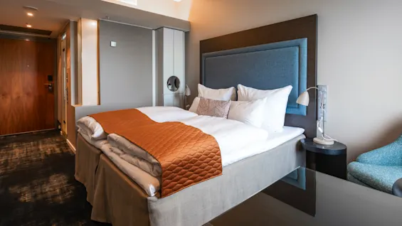 A neatly arranged bed with white linens and an orange quilt lies in a modern hotel room. Nearby, a turquoise chair and dark wooden door complement the neutral-toned decor.