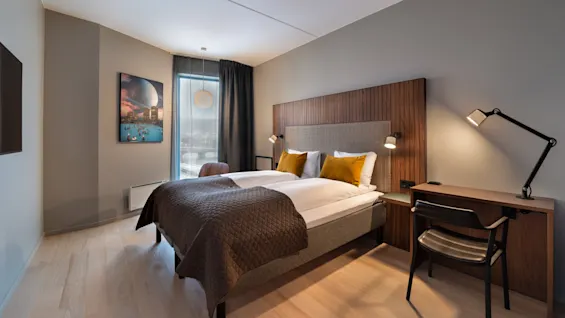 Et pent arrangert hotellrom med dobbelseng og gule puter, en trehodegjerde, et maleri på veggen, og et vindu med gardiner. Et skrivebord med lampe står i nærheten.
