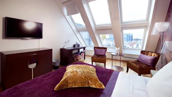 Et koselig loftsoverom med en myk lilla seng, stilige møbler, og takvinduer som ser ut over bysilhuetten.