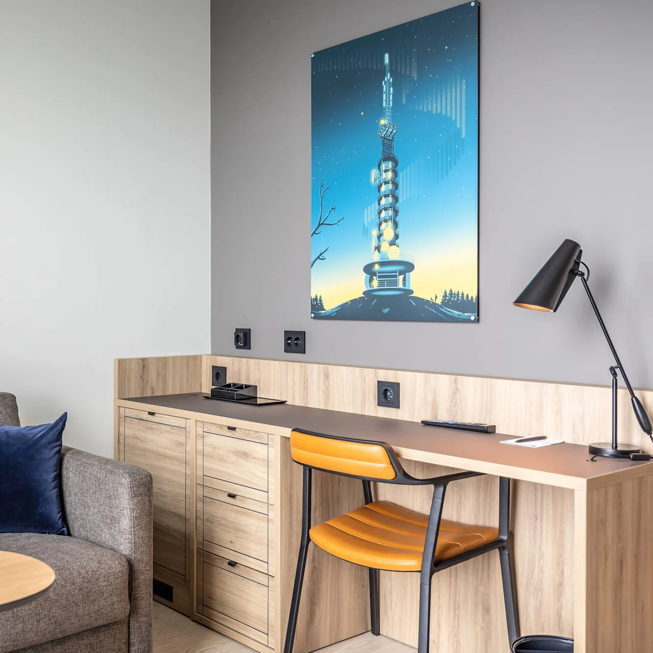 A gray fabric sofa with a blue cushion is beside a wooden desk, featuring a black lamp and a chair, in a room with a poster of a tower.