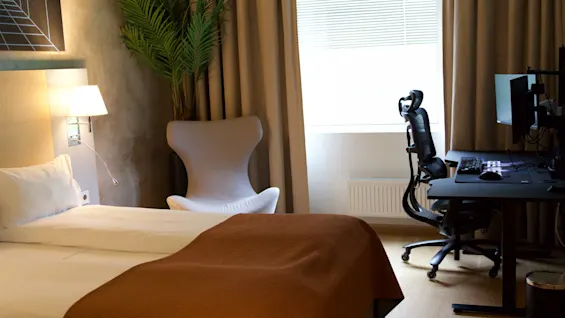 Moderne hotelværelse med en opredt seng og et dedikeret gamingområde med computer og ergonomisk stol.