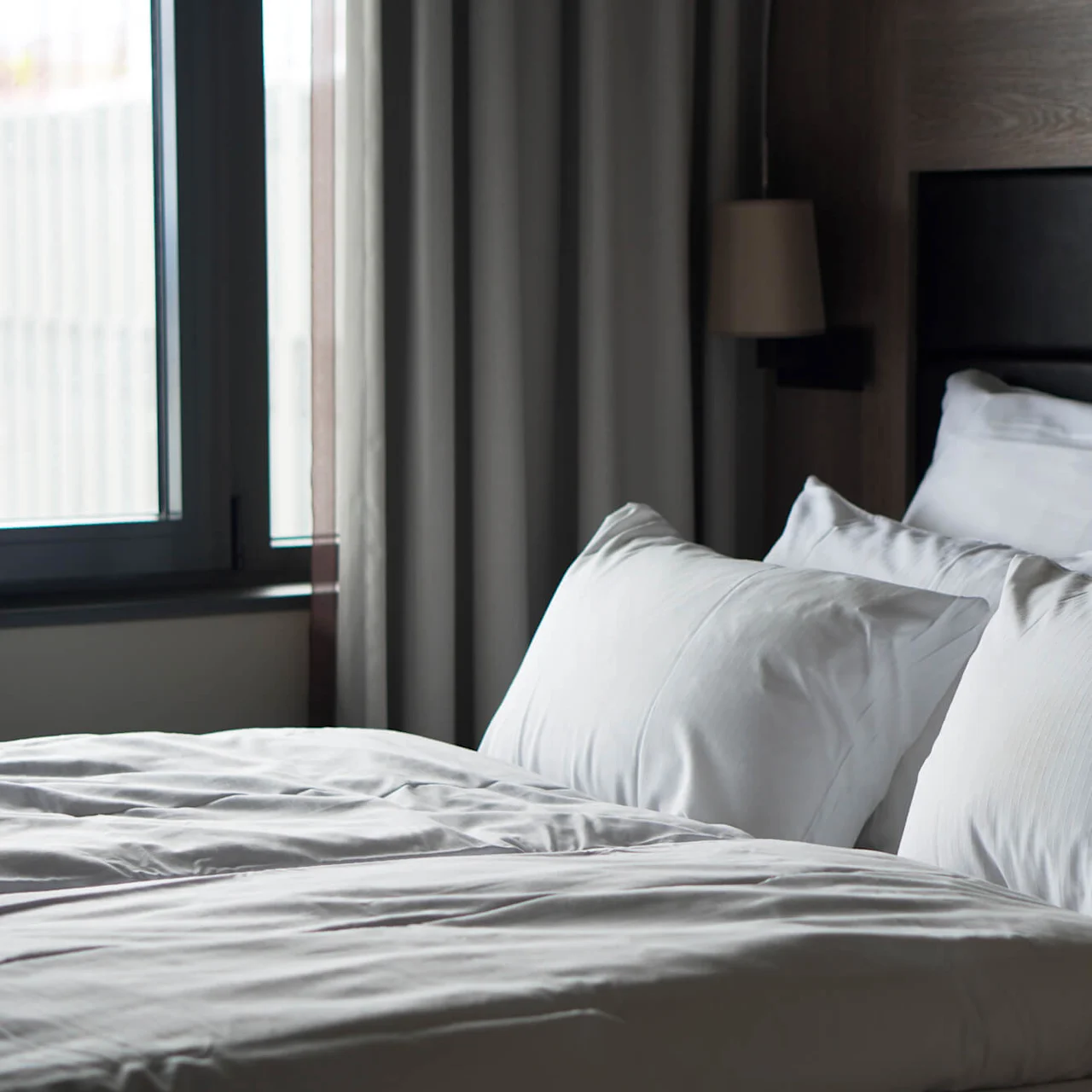 A neatly made bed with white pillows and duvet against a wooden headboard, situated near a window with closed curtains in a softly lit bedroom.