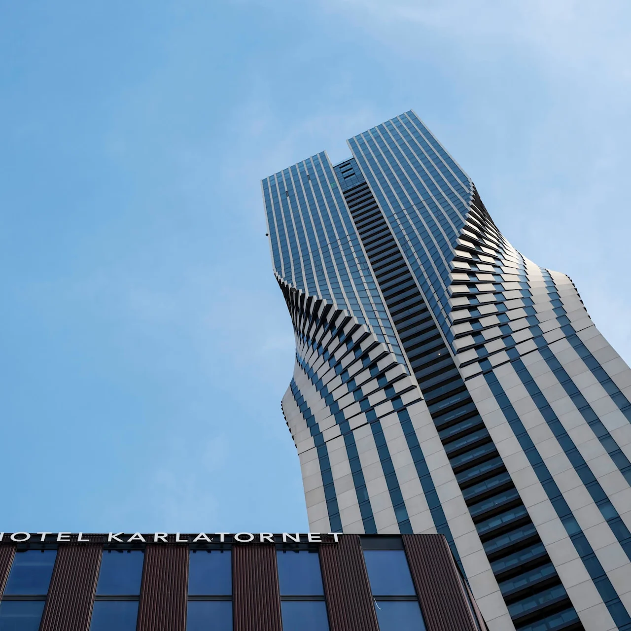 En vriden skyskrapa sträcker sig uppåt mot en klarblå himmel. Nedanför visar en modern byggnadsfasad texten "HOTEL KARLATORNET".