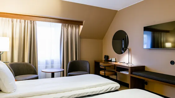 Et hotelværelse har to pænt redte senge. Ved siden af, en lille siddeplads med to stole og et rundt bord ved et gardindækket vindue. Overfor, et vægmonteret TV og et skrivebord med spejl og stol.