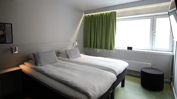 To pent oppredde enkeltsenger med mønstrede puter er i et minimalistisk rom. Et stort vindu med grønne gardiner gir naturlig lys. En liten rund puff er nær radiatoren.