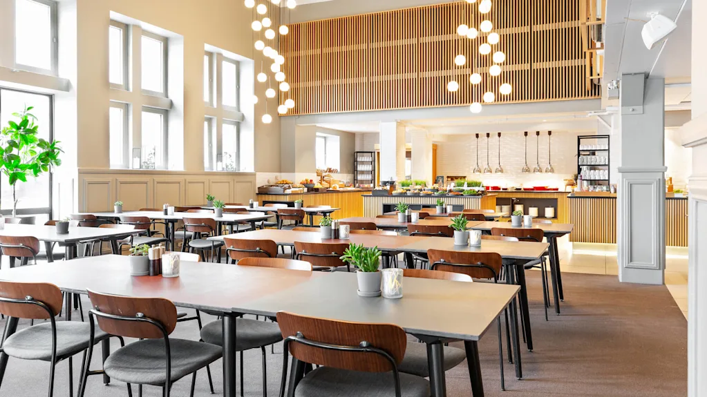 Bord med træstole arrangeret i et rummeligt, moderne spiseområde. Hængelamper hænger over og oplyser dekorative planter på bordene. En buffet med forskellige madvarer er synlig i baggrunden.