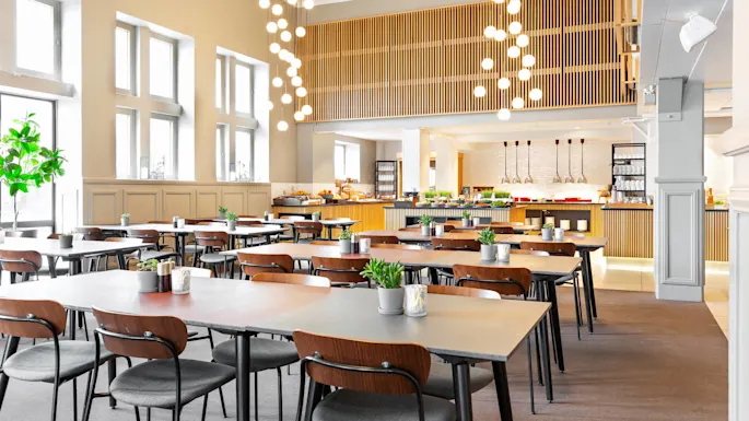 Pöydät puutarhoineen järjestetty tilavaan, moderniin ruokatilaan. Riippuvat valaisimet valaisevat pöydillä olevia koristekasveja. Taustalla näkyy buffet erilaisine ruokalajeineen.