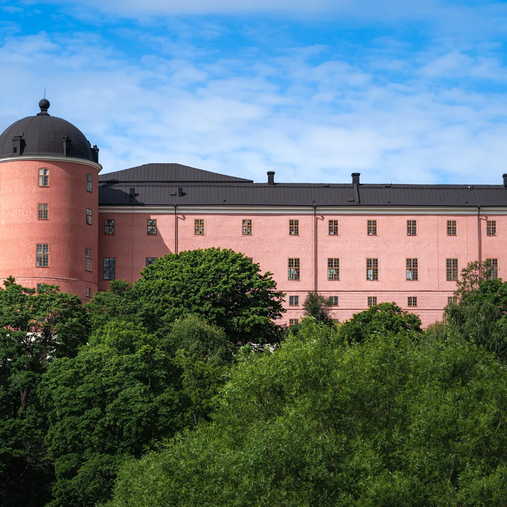 Uppsala linna, suuri vaaleanpunainen rakennus pyöreällä tornilla, kurkistaa esiin rehevien vihreiden puiden takaa sinisen taivaan alla.