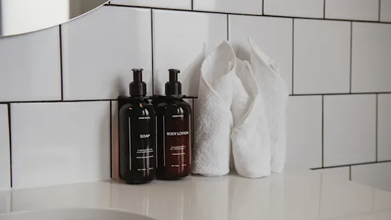 Flaskor märkta "SOAP" och "BODY LOTION" på en vit badrumshylla, placerade bredvid vikta vita handdukar, mot en vit kakelvägg.