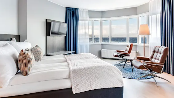 Et pænt indrettet hotelværelse med en seng med mønstrede puder, to brune stole vendt mod en udsigt over havet gennem store vinduer, blå gardiner og et tv på væggen.