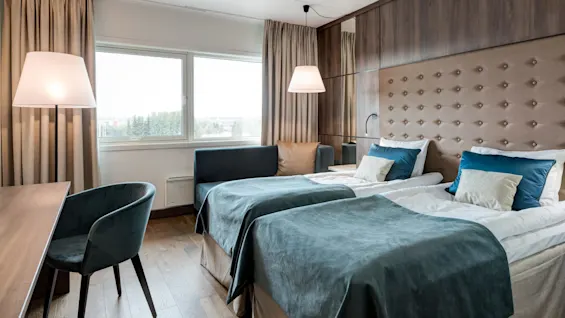 Two neatly made twin beds with blue bedding and pillows, positioned in a well-lit room with wooden floors, a padded headboard, and a large window showing a view of trees.
