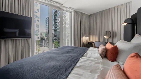 A neatly made bed with orange cushions faces a wall-mounted TV; tall windows reveal modern buildings outside. A desk with a lamp and mirror is on the right.  