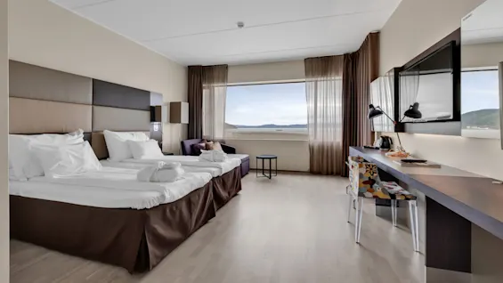 Bed positioned near a large window displaying a scenic view. Adjacent lounge chair, table, and desk with lamp and chair. Room features neutral colors and natural light.