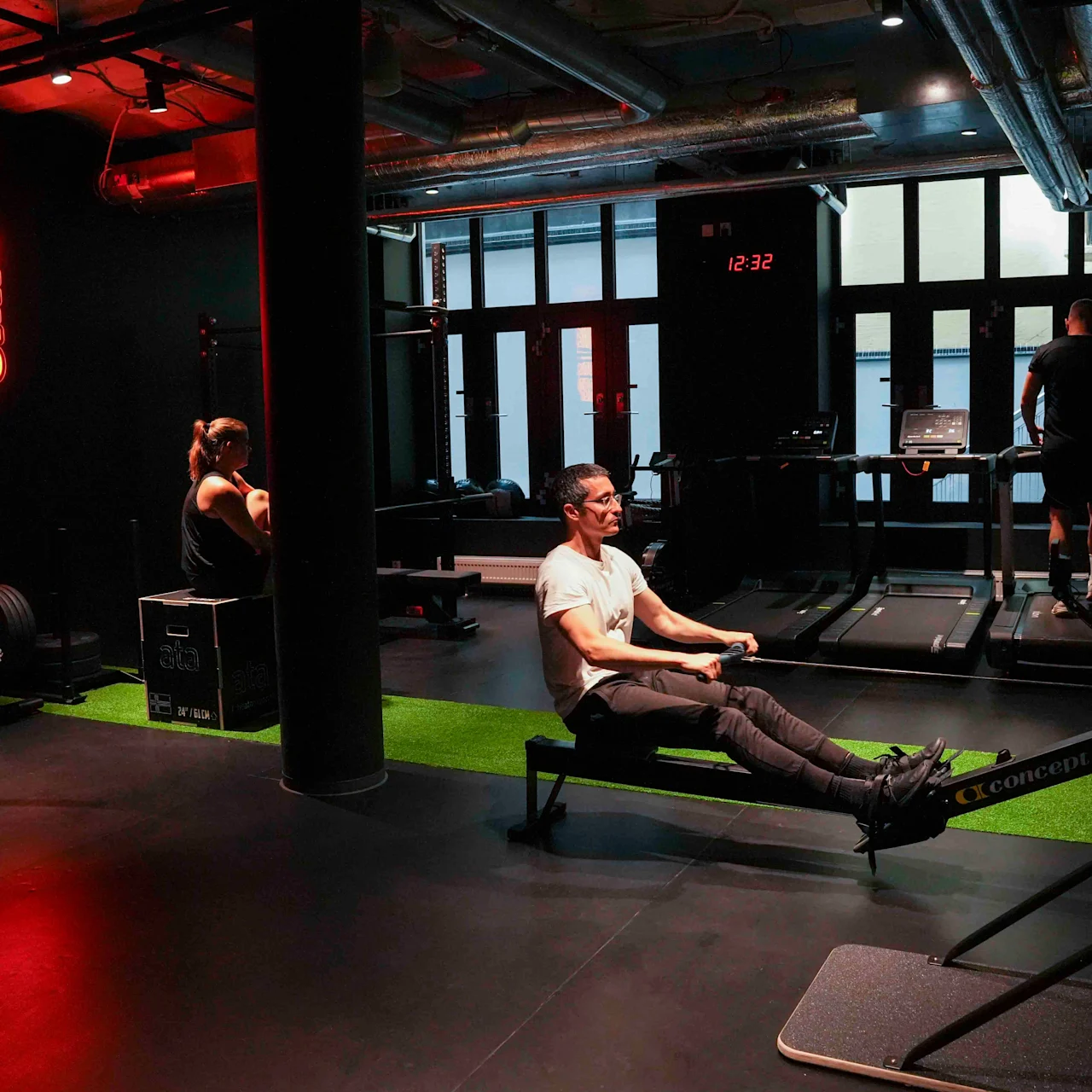 En person ror på en robåtmaskin på et treningssenter, under et neonskilt med teksten "ONE MORE REP". To andre personer bruker treningsutstyr i bakgrunnen, i svak belysning.