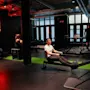 En person ror på en robåtmaskin på et treningssenter, under et neonskilt med teksten "ONE MORE REP". To andre personer bruker treningsutstyr i bakgrunnen, i svak belysning.