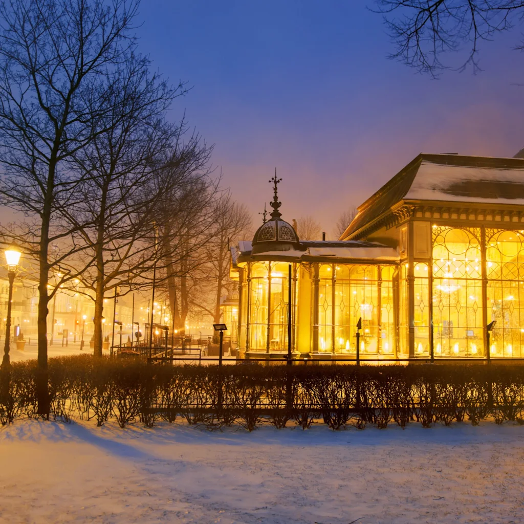 Restaurant Kappeli Helsinki naturally, a warmly lit building in a snowy park at dusk, creating a cozy winter atmosphere.