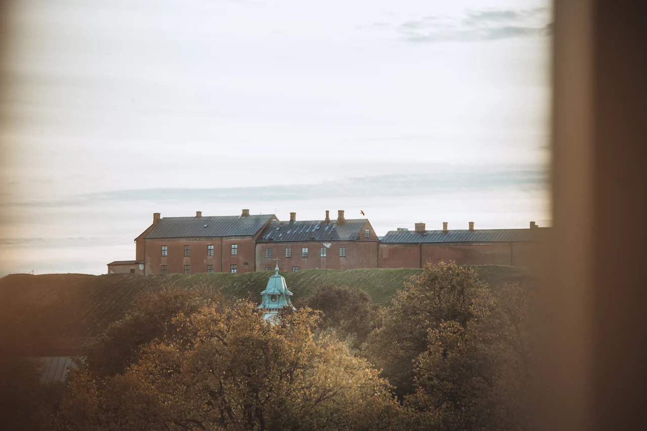 A scenic view of a grand brick building atop a grassy hill, framed by autumn trees and a window, under a soft sky.