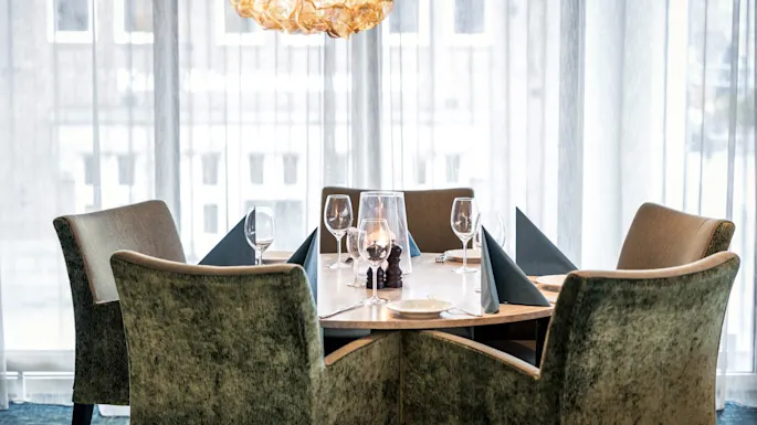 Pyöreä ruokapöytä on katettu laseilla ja lautasliinoilla, ympärillä pehmustetut tuolit. Koristeellinen riippuvalaisin roikkuu yllä, taustalla ohuet verhot ja rakennuksia.