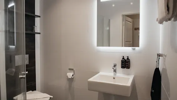 Et moderne og rent badeværelse med en vask, et oplyst spejl og et toilet. En sort taske med Lve hænger på væggen.