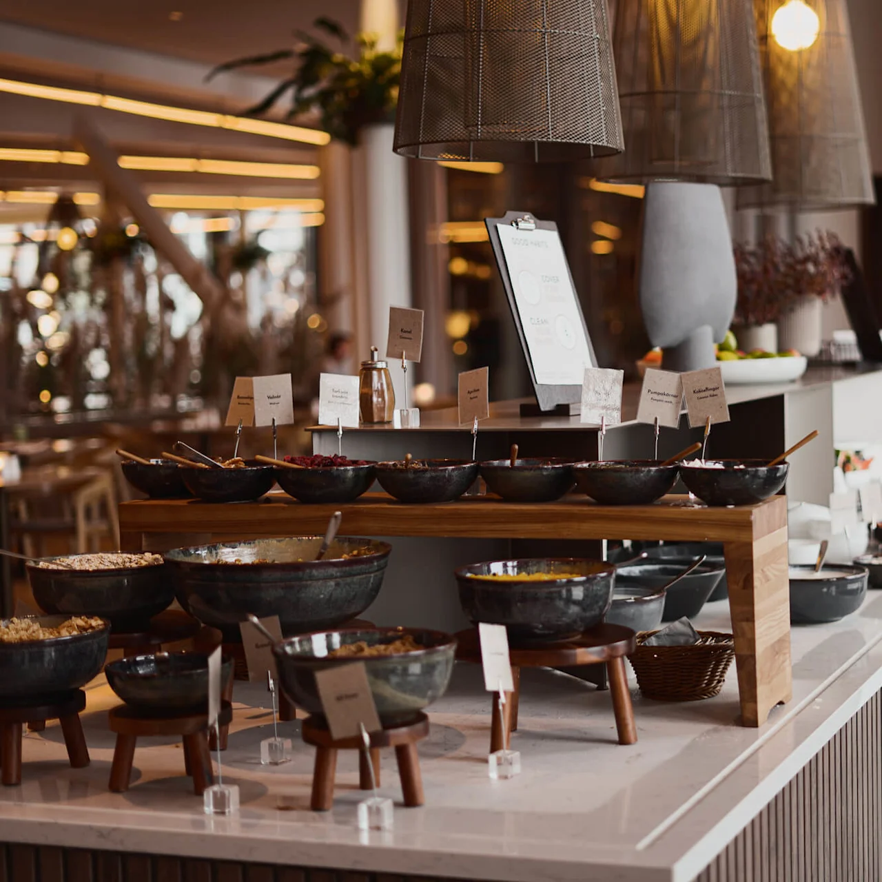 Et buffébord med forskjellige merkede skåler med mat i en moderne restaurant, omgitt av stemningsbelysning og dekorative planter. Synlige skilt inkluderer "Nötter," "Honung," "Yoghurt," og "Frön."