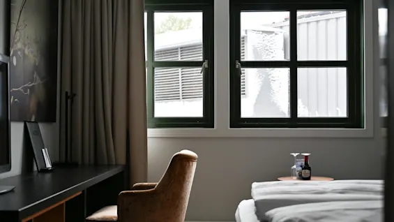 En mysig hotellrum har en brun stol och skrivbord vid ett gardinbeklätt fönster med utsikt över snö. Ett litet bord bredvid sängen har vin och glas.
