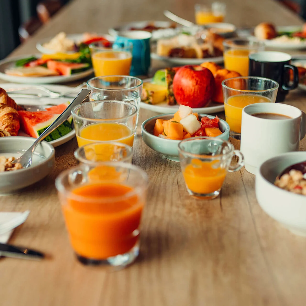 A breakfast table with various foods including croissants, fruit, cereals, and multiple glasses of orange juice, creating a vibrant and inviting spread.