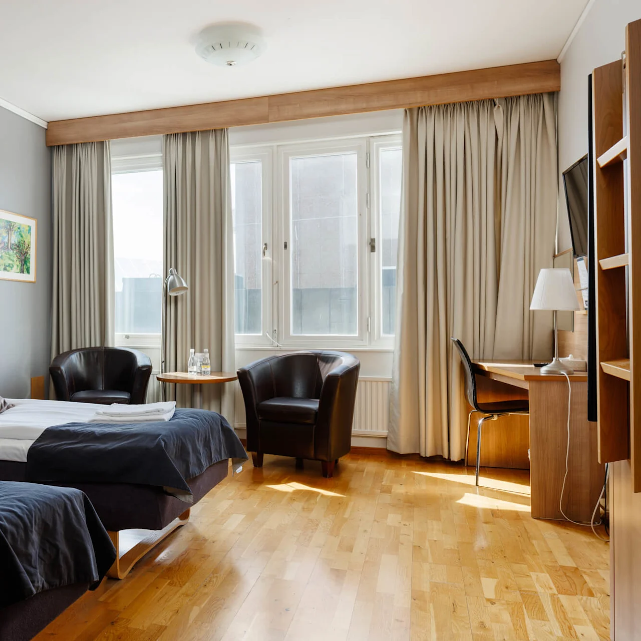 Två prydligt bäddade enkelsängar med lampor på sidobord, placerade i ett hotellrum med trägolv och stora fönster. Stolar och ett litet bord står vid fönstret.