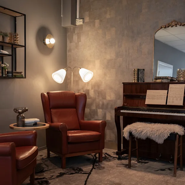 An inviting lounge at Home Hotel Grand Bodø featuring comfortable leather armchairs, a piano with sheet music, and warm lighting.