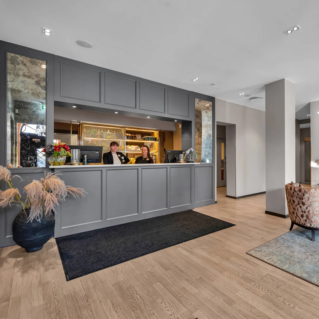 Två personer står bakom en receptionsdisk på ett hotell, leende och utför arbetsuppgifter i en modern dekorerad lobby med mysigt sittområde.