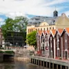 Röda trähus står vid en flodkant och speglas i lugnt vatten. Bakom dem finns historiska stenbyggnader och lummiga träd under en klarblå himmel med några få moln.