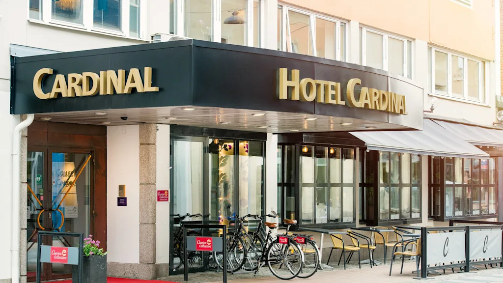 A hotel entrance with a black and gold sign reading "Cardinal Hotel Cardinal." Bicycles are parked and chairs arranged outside on a sidewalk.