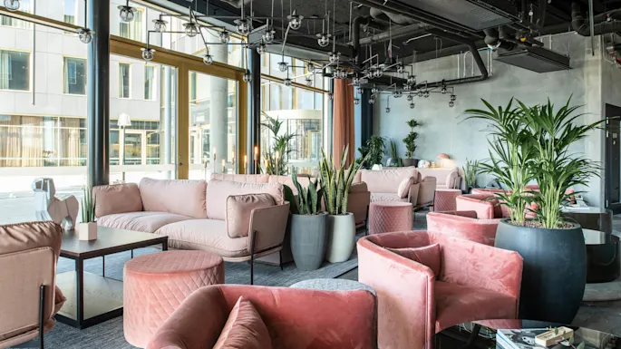 Luksuriøs lounge med rosa fløyelsofaer og stoler arrangert rundt lave bord. Store potteplanter tilfører grønt, i et moderne interiør med gulv-til-tak-vinduer og dekorativt tak.