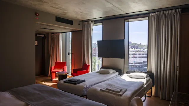 To pænt redte senge i et moderne hotelværelse vender mod et vægmonteret TV. Store vinduer med byudsigt er omgivet af lette gardiner. Røde stole og et bord står nær vinduet.