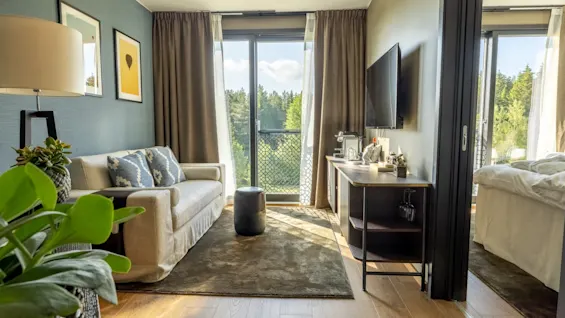 Ett mysigt vardagsrum med en beige soffa med mönstrade kuddar, vänd mot ett stort fönster med gardiner; en TV och ett bord står till höger, med skogsutsikt utanför.