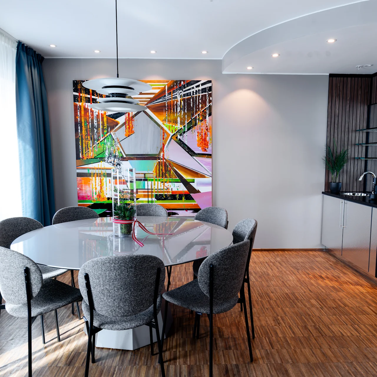 Dining table surrounded by gray chairs, set beneath a modern pendant light. In the background, a vibrant abstract painting hangs on a light gray wall, near a kitchenette and a window with curtains.