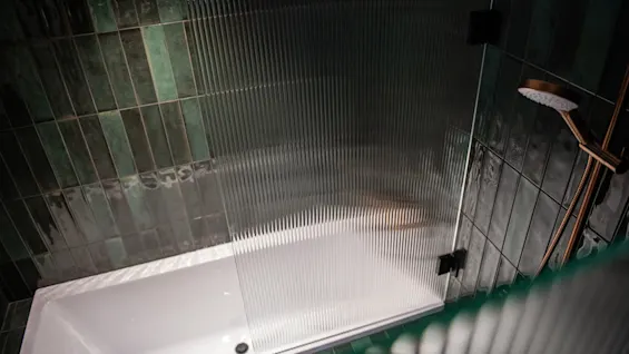 Ett ribbat glasduschskärms står bredvid ett tomt vitt badkar, omgivet av mörkgröna kaklade väggar. Ett bronsfärgat duschhuvud är monterat till höger.