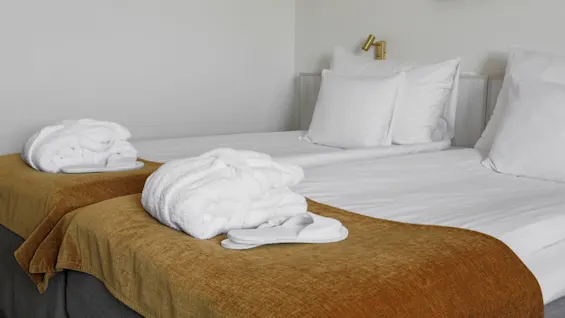 A neatly made bed with white sheets and pillows holds folded bathrobes and slippers on brown blankets. The setting is a minimalistic room with a white wall.