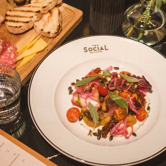 En anrettet gourmet-forrett på en tallerken ved siden av et trebrett med spekemat, ost og grillet brød.