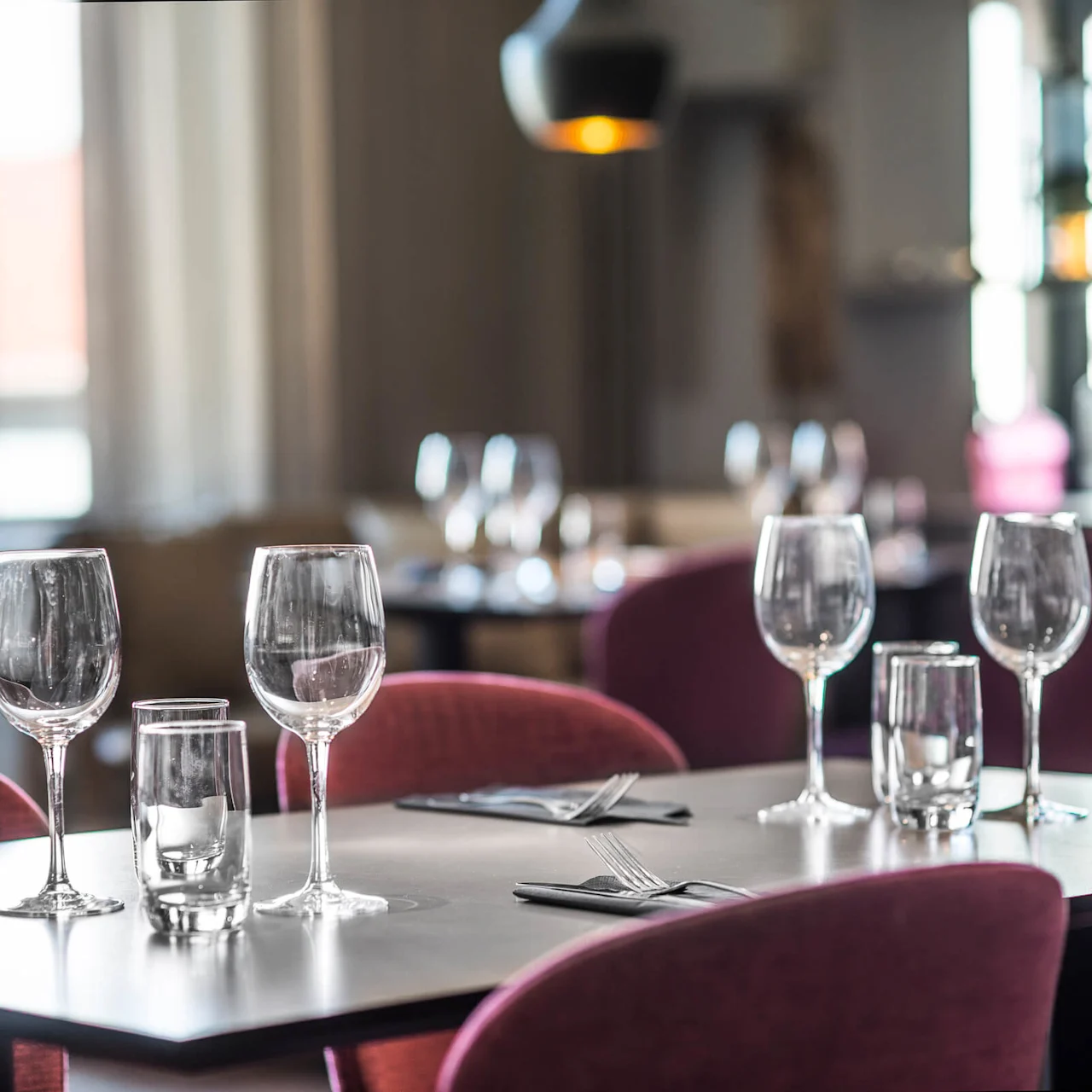 Matbord dukat med tomma vinglas och servettinlindade bestick, omgiven av röda stolar i en mjukt upplyst restaurang. Ingen text finns.