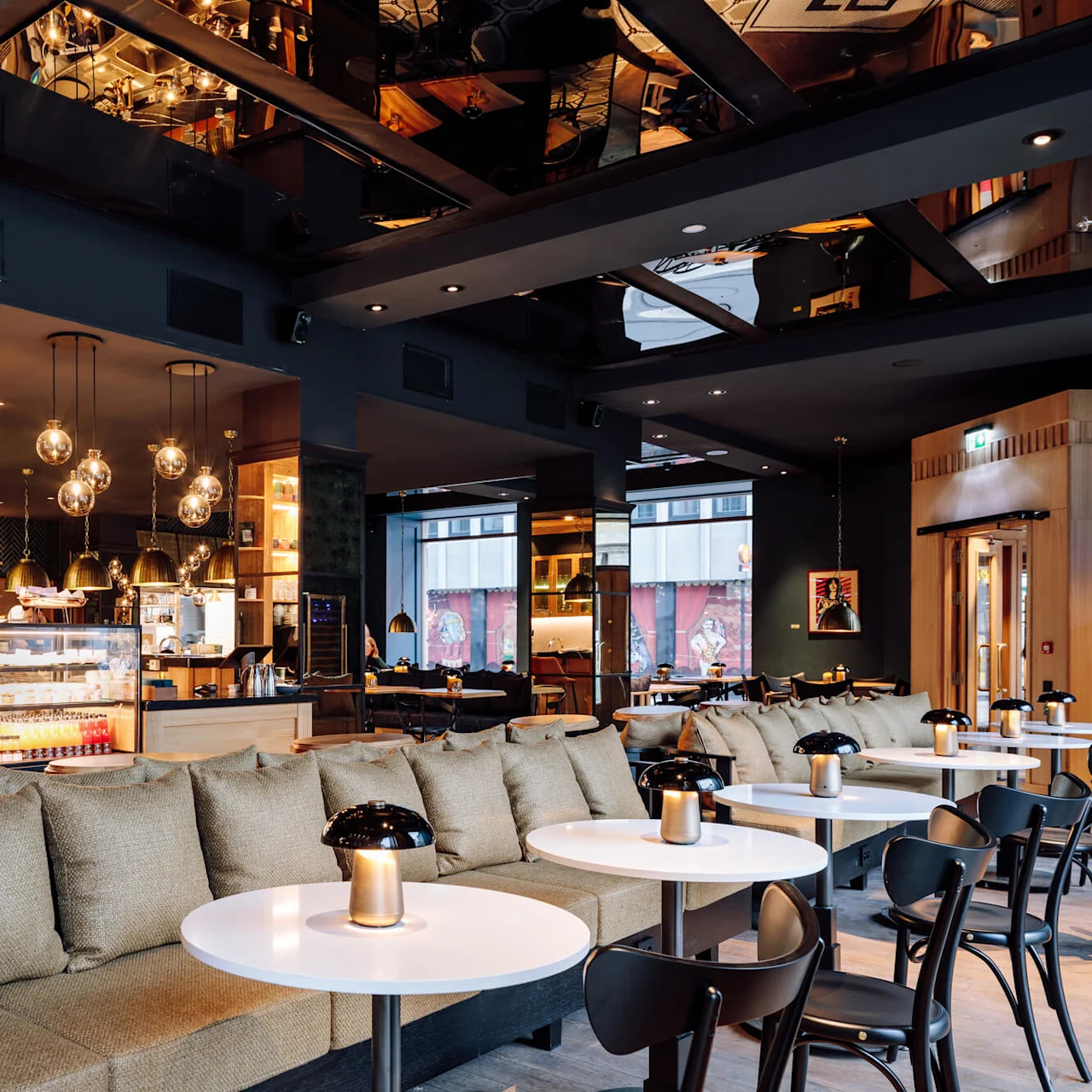 Café interior, featuring a cushioned sofa with white tables and lamps, surrounded by chairs. Pastry and beverage display counter under hanging globe lights; large windows with a street view.