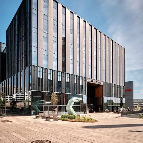 En moderne glassbygning med vertikale paneler står på en brolagt gårdsplass med benker og en metallskulptur. Skilt viser, "Quality Hotel Hasle Linie."