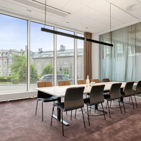 Et lang bord med ti stoler er plassert i et moderne møterom. Store vinduer viser utsikt over en historisk bygning utenfor. Minimalistisk dekor øker den romslige følelsen.