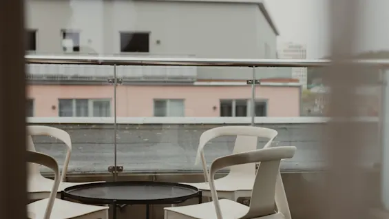 Four white chairs surround a round black table, situated on a balcony with glass railing, overlooking distant, blurred buildings under a cloudy sky.