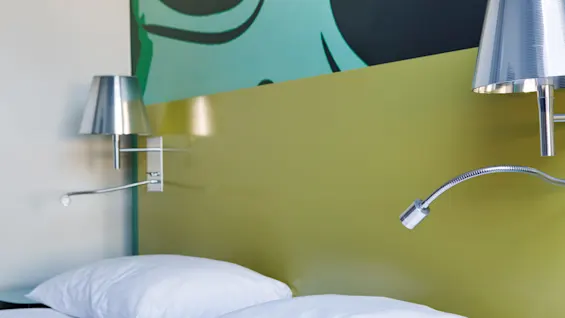 White pillows rest on a bed beneath two metallic reading lamps, against a green headboard with abstract artwork above.