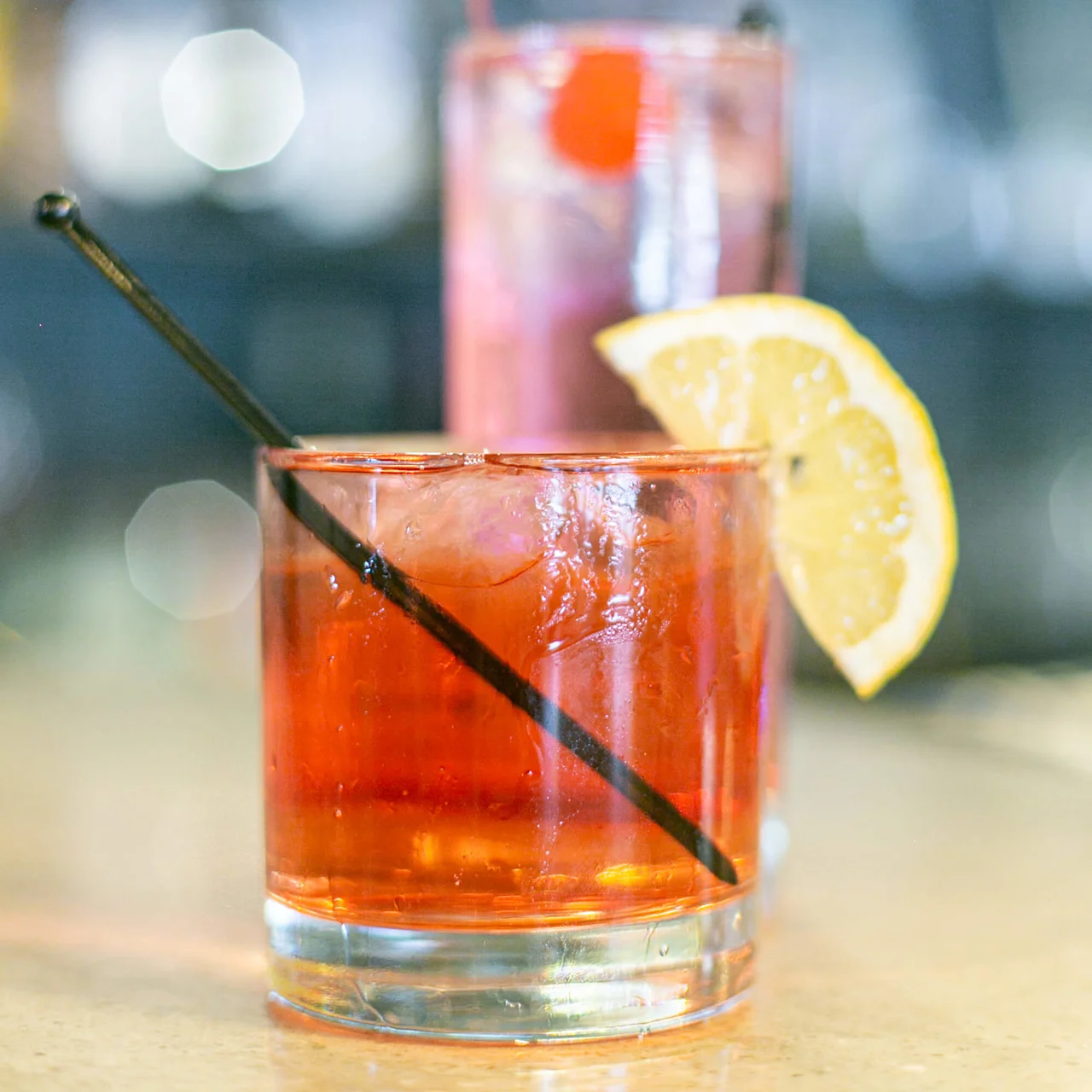 En rød cocktail med is og en citronskive på kanten står i et lavt glas med et sort sugerør, mod en sløret bardisk.