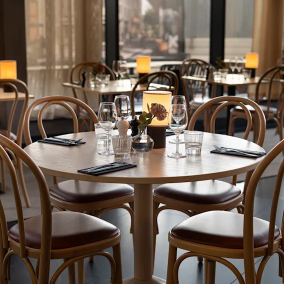 Round wooden dining table set with glassware, napkins, and flowers in a dimly lit restaurant with curved chairs and illuminated ambient candles.